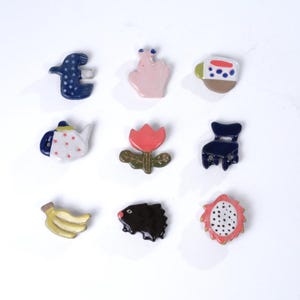 May include: Nine ceramic pins in assorted shapes and colours. The pins depict a blue bird with white spots, a pink hand, a mug, a teapot, a red tulip, a blue chair, a banana, a black hedgehog, and a dragon fruit slice.