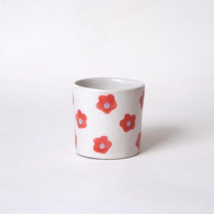 Könnte beinhalten: Eine weiße Keramiktasse mit zylindrischer Form. Die Tasse ist mit einem sich wiederholenden Muster aus roten Blumen mit violetten Zentren verziert. Die Tasse eignet sich zum Halten von Getränken und ist ein dekorativer Gegenstand.