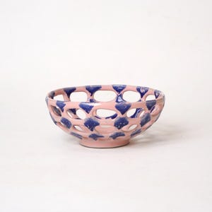 Puede incluir: Un cuenco de cerámica rosa con un diseño de celosía y detalles azules. El cuenco tiene un patrón calado con aberturas ovaladas. El interior y el exterior son rosas, con patrones azules en forma de diamante.