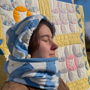 Peut inclure: Un capuchon et un tour de cou matelassés à carreaux bleus et blancs. La personne porte une veste marron. Une courtepointe vintage avec des motifs floraux jaunes, roses et bleus est suspendue en arrière-plan.