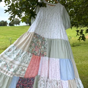 May include: A patchwork maxi dress featuring floral, gingham, and solid fabric panels in white, pink, green, and blue. The dress has short sleeves and a tiered skirt, hanging on a wooden hanger.