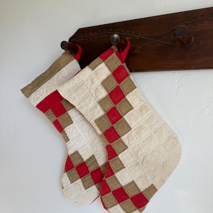 Puede incluir: Dos calcetines navideños acolchados con un diseño de patchwork de cuadrados de tela en color crema, tostado y rojo. Cada calcetín tiene una pequeña trabilla de tela roja en la parte superior para colgar, con costuras visibles en forma de rejilla.