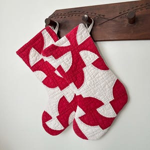 May include: Two quilted Christmas stockings featuring a geometric pattern of interlocking red and white curved shapes. Each stocking has a small white fabric loop at the top for hanging, showcasing detailed stitching throughout the fabric surface.