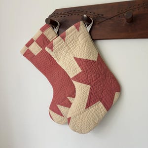 May include: Two quilted Christmas stockings featuring a geometric star pattern in muted red and cream fabric. Each stocking has a small white fabric loop at the top for hanging, with visible stitching texture across the entire surface of the fabric.
