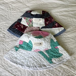 May include: Two bucket hats made from quilted fabric. The top hat features a patchwork design with dark blue, white, and floral patterns. The bottom hat is white with green and pink floral shapes. Both hats have a Northern Homemakers brand label.