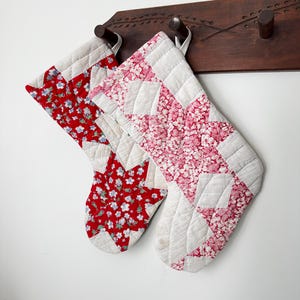 Puede incluir: Dos calcetines navideños acolchados colgados de un perchero de madera. El calcetín izquierdo presenta una tela floral roja con patrones de estrellas blancas. El derecho presenta una tela floral rosa con patrones de estrellas blancas.