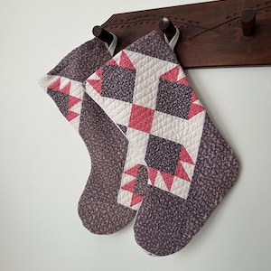 May include: Two quilted Christmas stockings featuring a geometric patchwork design. The fabric patterns include dark purple floral prints, solid white sections, and bright pink triangular accents. Each stocking has a small white loop for hanging.