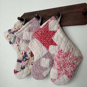 May include: Three quilted Christmas stockings hanging from a dark brown wooden rack. Each stocking features a unique patchwork design with various red, white, and floral fabric patterns. The stockings are constructed with visible stitching.