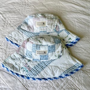 Puede incluir: Dos sombreros de pescador confeccionados con tela de patchwork acolchada en tonos blanco, azul y rosa suave. Cada sombrero presenta una etiqueta blanca rectangular en el frente con el texto Northern Homemakers y un icono de ave, con borde de cuadros vichy azul.
