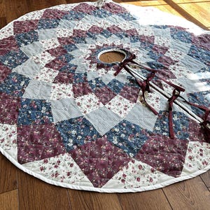 May include: A circular quilted tree skirt featuring a patchwork design of burgundy, navy blue, and white floral and gingham fabrics. The skirt has a central circular opening, a white piped edge, and three sets of burgundy ribbon ties for closure.