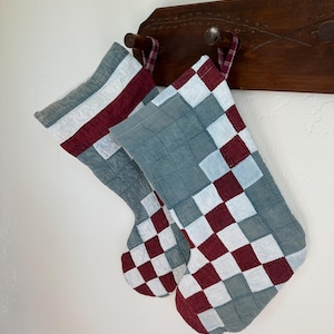 Könnte beinhalten: Zwei gesteppte Weihnachtsstrümpfe mit einem Patchwork-Design aus salbeigrünen, dunkelburgunderroten und weißen Stoffquadraten. Jeder Strumpf hat eine kleine Stoffschlaufe zum Aufhängen und ein sichtbares, gestepptes Gittermuster.