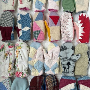 Puede incluir: Una colección de mitones acolchados en varios colores y patrones. Los mitones presentan diseños de patchwork, estampados florales y colores lisos. Algunos tienen forros rosas o azules. Los mitones están dispuestos sobre una superficie blanca.