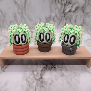 Puede incluir: Tres pequeñas figuritas de plantas verdes con caras y macetas de diferentes colores. Las macetas son marrones, naranjas y grises. Las figuritas están en un estante de madera.