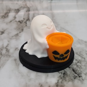 May include: A white ghost figurine holding a small orange pumpkin-shaped container with a black jack-o'-lantern face. The figurine is sitting on a black circular base.