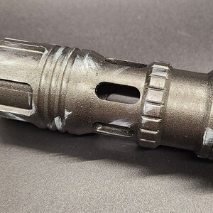 Pu&ograve; includere: Primo piano di un'impugnatura di spada laser in metallo nero con un design di vernice bianca. L'impugnatura ha una superficie testurizzata e un piccolo foro sul lato.