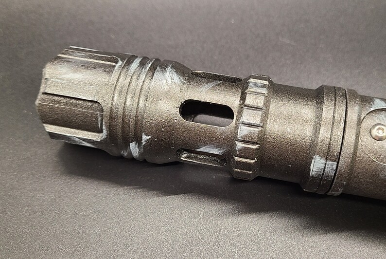 Pu&ograve; includere: Primo piano di un'impugnatura di spada laser in metallo nero con un design di vernice bianca. L'impugnatura ha una superficie testurizzata e un piccolo foro sul lato.