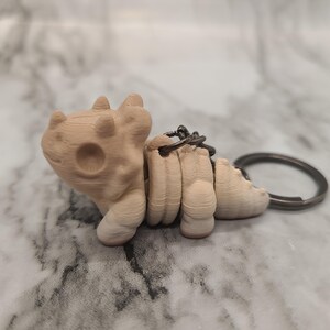 Puede incluir: Un llavero beige impreso en 3D con forma de criatura estilizada. El llavero tiene un llavero circular y una cadena que conecta el cuerpo de la criatura. La criatura tiene una cabeza redondeada con tres pequeñas protuberancias y una gran hendidura circular.