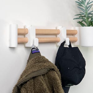 May include: A wall-mounted coat rack featuring two horizontal light wood dowels held by white plastic brackets. The rack includes two white hooks holding an olive green fleece jacket and a black baseball cap, plus a white planter on the right.