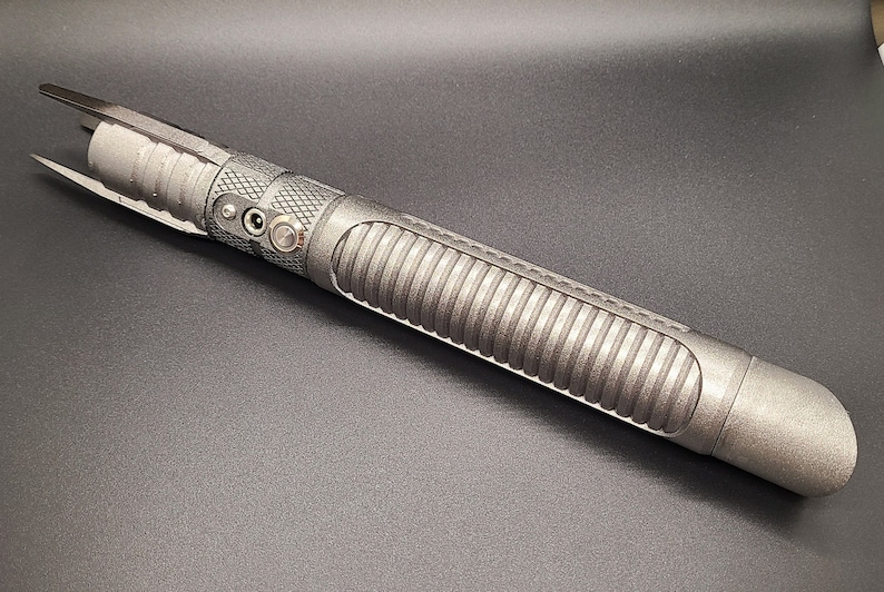 May include: A silver lightsaber hilt with a textured grip and a black button. The hilt has a rounded end and a pointed tip.