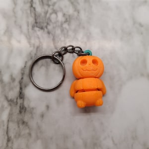 May include: Orange pumpkin-shaped keychain with a carved face and a teal string loop. The keychain is attached to a silver metal key ring.