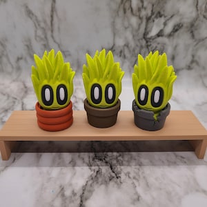 Puede incluir: Tres pequeñas figuras verdes con forma de planta, con caras y ojos negros, están en macetas de diferentes colores. Las macetas son marrones, naranjas y grises. Las figuras están en un estante de madera.