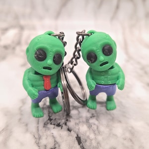 May include: Two green alien keyrings. Each alien figure has large black eyes, a small mouth, and is wearing blue shorts. One alien has a red tie. The keyrings are attached to a silver chain and a split ring.