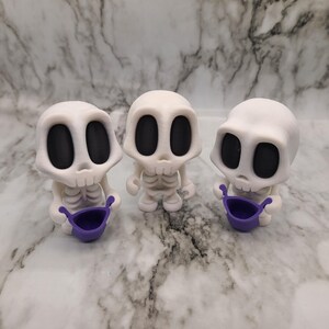 May include: Three white skeleton figurines with large black eye sockets and rib cages. Each figurine holds a small purple bucket. The figures are standing on a marble-patterned surface.