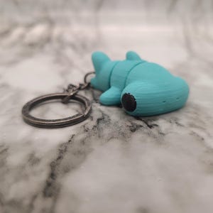 May include: A turquoise 3D-printed keychain featuring a stylized animal figure with a black accent on its rear. The keychain has a silver-colored ring attached. The object is resting on a marbled surface.