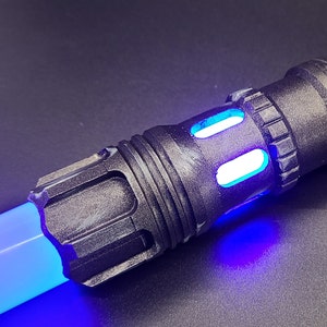 Pu&ograve; includere: Primo piano di un'impugnatura di spada laser nera con una lama di spada laser blu. L'impugnatura ha una superficie testurizzata e una luce blu che emana dall'estremit&agrave;.