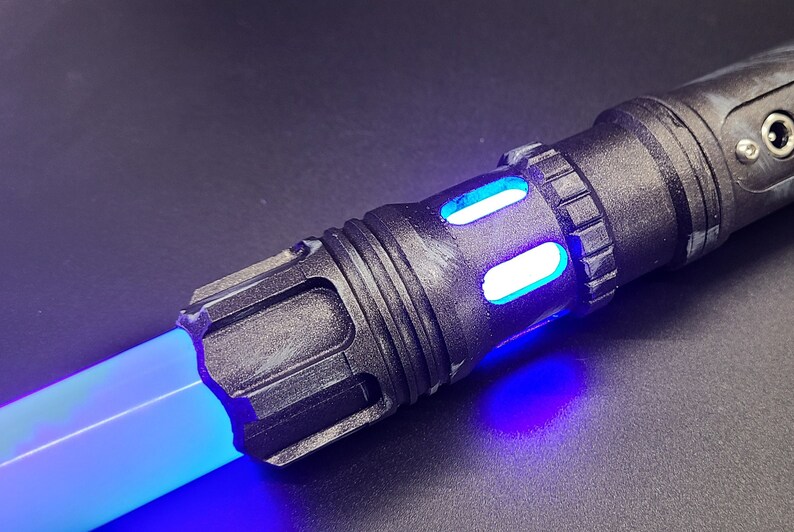 Pu&ograve; includere: Primo piano di un'impugnatura di spada laser nera con una lama di spada laser blu. L'impugnatura ha una superficie testurizzata e una luce blu che emana dall'estremit&agrave;.