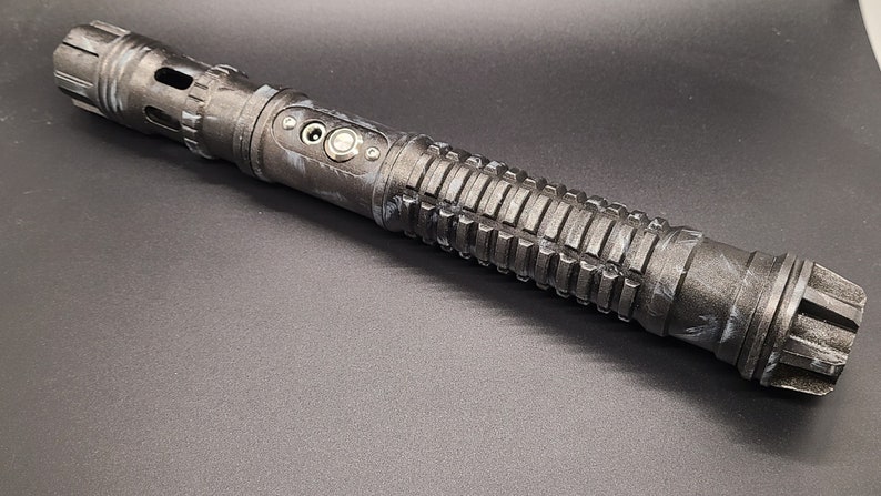 Pu&ograve; includere: Una spada laser nera e argentata con un'impugnatura testurizzata e un pulsante argentato. La spada laser ha un motivo di mimetizzazione nero e bianco.