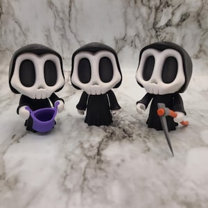 May include: Three figurines of the Grim Reaper, each with a black hooded robe and a white skull face with large eye sockets. One holds a purple bucket, and another holds a scythe with an orange handle. The figurines are standing on a marble surface.