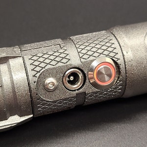 May include: Close-up of a black metal object with a power adapter and a red-ringed button. The object has a textured surface.