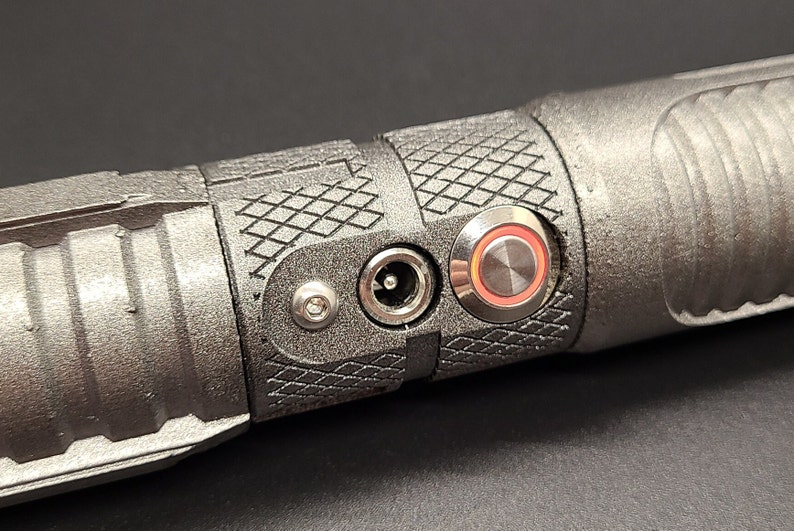 May include: Close-up of a black metal object with a power adapter and a red-ringed button. The object has a textured surface.