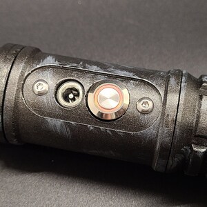 Pu&ograve; includere: Un'impugnatura di spada laser nera con un pulsante argentato e una porta di alimentazione. L'impugnatura ha una superficie testurizzata e una vite argentata sul lato.