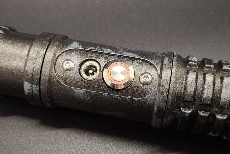 Pu&ograve; includere: Un'impugnatura di spada laser nera con un pulsante argentato e una porta di alimentazione. L'impugnatura ha una superficie testurizzata e una vite argentata sul lato.