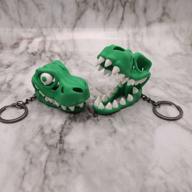 May include: Two green dinosaur skull keychains with white teeth. One keychain is open-mouthed and the other is closed-mouthed.