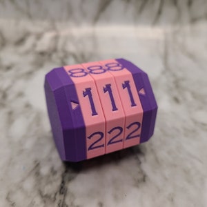 May include: A purple and pink hexagonal-shaped counter with three rotating sides. Each side has a different number printed on it: 888, 111, and 222.