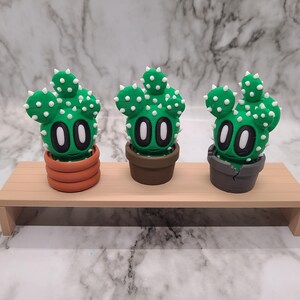 May include: Three small green cactus figurines with white dots and black eyes, each in a different colored pot. The pots are brown, orange, and gray.