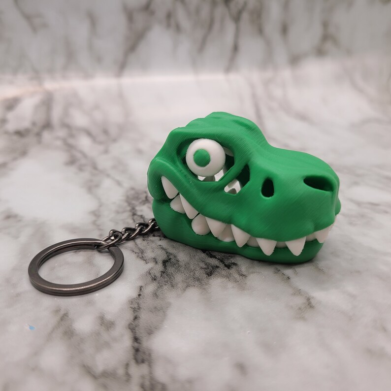 May include: A green plastic dinosaur skull keychain with a white eye and teeth. The keychain is attached to a silver metal ring.