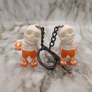 Peut inclure: Deux porte-clés squelettes blancs portant des shorts de bain orange et tenant un ballon de plage. Les porte-clés sont attachés à une chaîne argentée.
