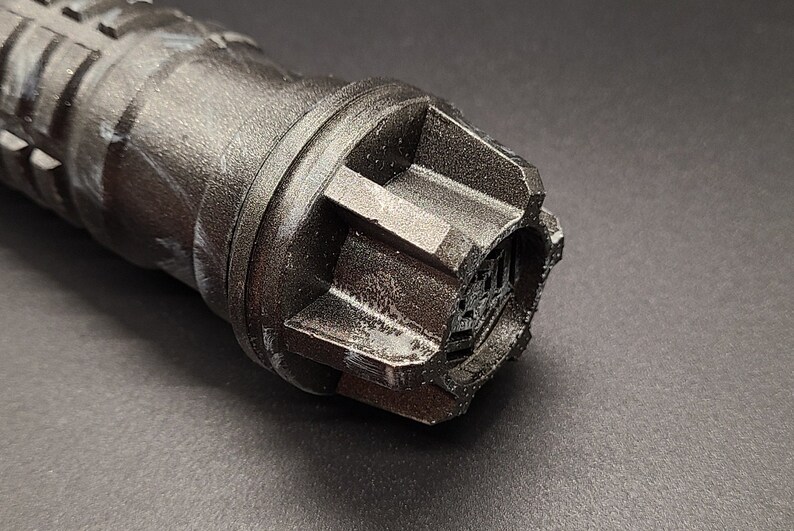 Pu&ograve; includere: Primo piano di un'impugnatura di spada laser nera con una superficie testurizzata e marmorea. L'impugnatura ha un tappo esagonale con un piccolo design intricato.