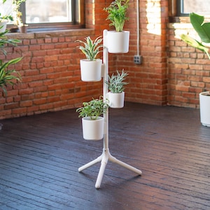 May include: A white vertical plant stand with a tripod base featuring four cylindrical white pots arranged at varying heights along a central pole. Each pot contains a different green houseplant, creating a tiered display of foliage.