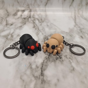 Peut inclure: Deux porte-clés araignées imprimés en 3D. L'un est noir avec des yeux rouges et l'autre est beige avec des yeux noirs.