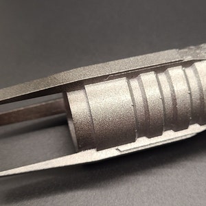 May include: A silver-colored lightsaber hilt with a textured surface and a black handle. The hilt has a unique design with a split end and a series of ridges.