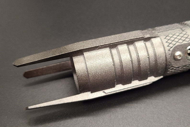 May include: A silver-colored lightsaber hilt with a textured surface and a black handle. The hilt has a unique design with a split end and a series of ridges.