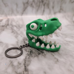 May include: A green dinosaur skull keychain with white teeth. The keychain has a silver metal ring.