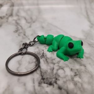 Peut inclure: Un porte-clés en forme de grenouille verte avec des yeux noirs. La grenouille est en plastique et a un corps segmenté. Le porte-clés est attaché à un anneau en métal argenté.