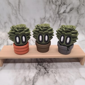 May include: Three small green succulent plants with white eyes and a smiling mouth are potted in different colored pots. The pots are orange, brown, and gray. The plants are sitting on a wooden shelf.
