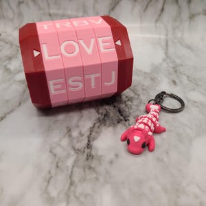 May include: A pink and red 3D printed keychain with a rotating cube that spells out "LOVE" and a pink lizard charm with white hearts.